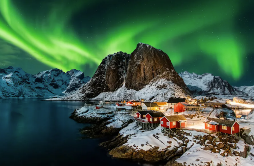 Green Northern Lights over red cabins and a snow-capped peak on the Norwegian coast